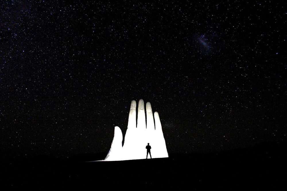 Mención honrosa 6: "Cuando Atacama con el cosmos se tocan, yo soy el testigo".  Autor: Carlos Gatica.  "La escultura Mano del Desierto, ubicada a 75 km. al sur de Antofagasta; sobre ésta, las Nubes de Magallanes". Foto tomada en marzo 2016; región de Antofagasta.